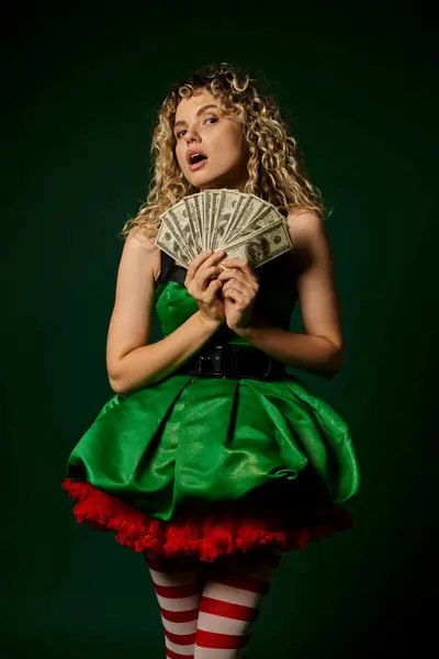 Astonished pretty new year elf in green dress and striped stockings holding cash looking at camera — Stock Photo