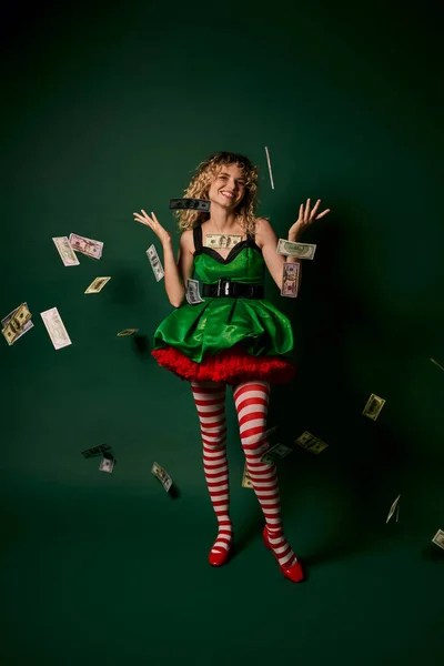 Mulher encaracolado alegre vestido como elfo do ano novo em pé sob chuva dinheiro alegremente olhando para a câmera — Fotografia de Stock