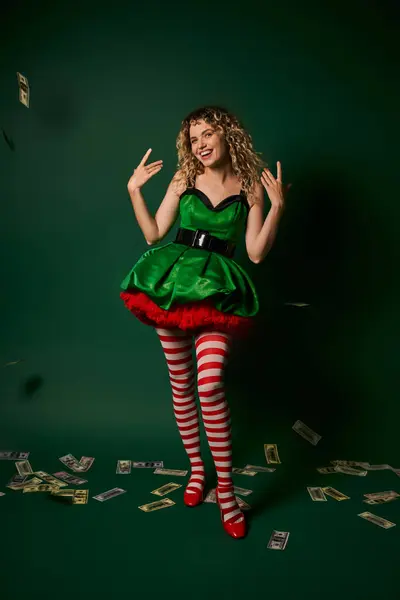 Smiley new year elf in green dress and stockings gesturing and smiling with cash on green floor — Stock Photo