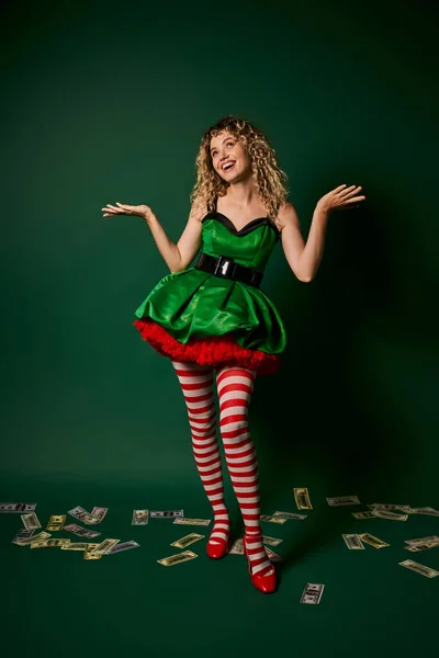Mujer alegre en vestido verde mirando hacia arriba y haciendo gestos con dinero en el suelo, concepto de año nuevo elfo - foto de stock
