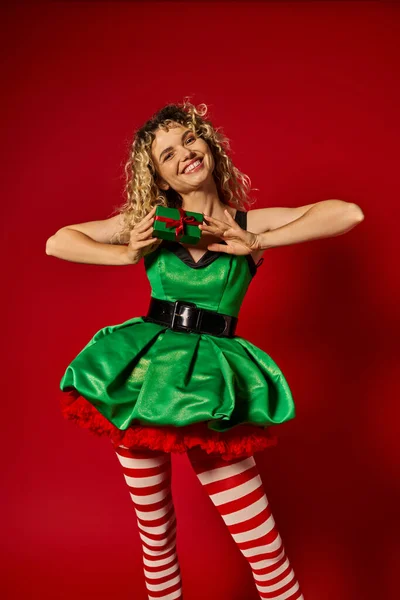 Alegre rizado mujer vestida como nuevo año elfo sonriendo sinceramente y mostrando presente en la cámara - foto de stock