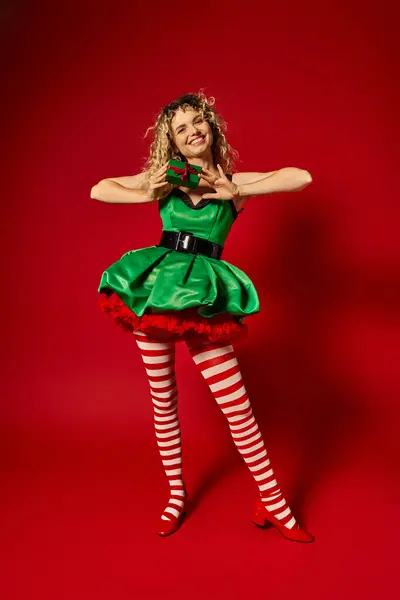 Atractiva mujer vestida como elfo año nuevo sonriendo y sosteniendo presente en la cámara en el telón de fondo rojo - foto de stock