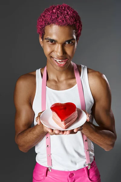Homme joyeux avec des cheveux roses montrant mini gâteau en forme de coeur et sortant la langue, la mode et le style — Photo de stock