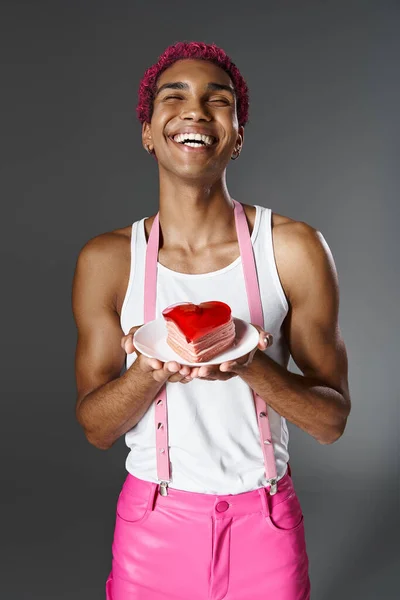 Joyeux jeune homme tenant mini gâteau en forme de coeur et souriant à la caméra, la mode et le style — Photo de stock