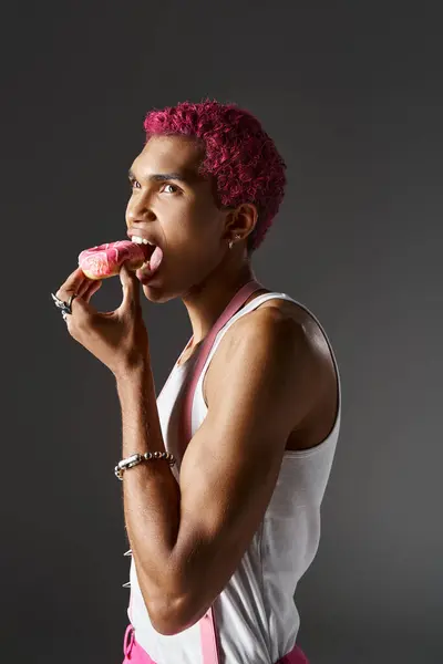 Beau modèle masculin avec des cheveux roses manger beignet posant sur fond gris, la mode et le style — Photo de stock