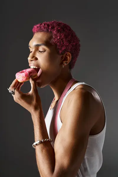 Bonito afro-americano homem com cabelo rosa e suspensórios comer saboroso donut, moda e estilo — Fotografia de Stock
