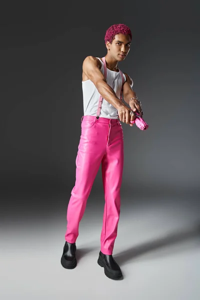 Hombre de moda con pelo rizado rosa con pantalones con tirantes y pistola de juguete rosa - foto de stock