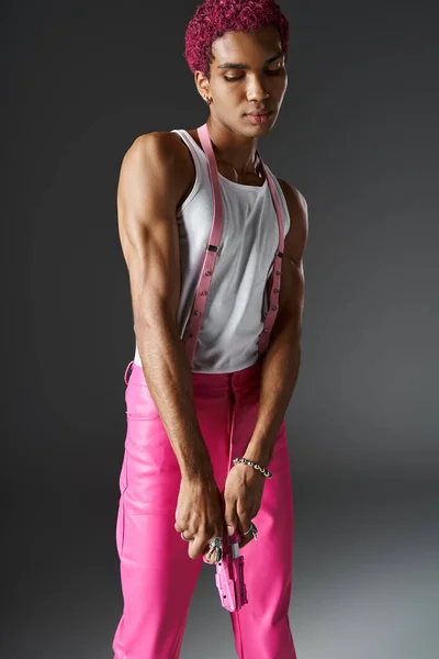 Hombre afroamericano elegante con el pelo rosa rizado mirando hacia abajo y posando con pistola de juguete rosa - foto de stock