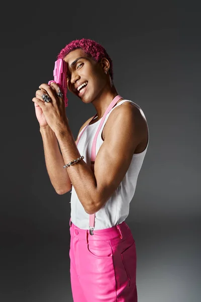 Beau gai homme avec les cheveux bouclés rose posant avec pistolet jouet rose et souriant à la caméra — Photo de stock