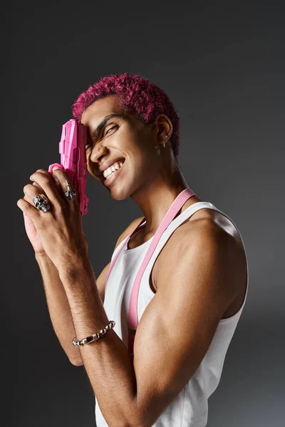 Modelo masculino alegre com cabelo rosa encaracolado posando em perfil com arma de brinquedo rosa e sorrindo para a câmera — Fotografia de Stock