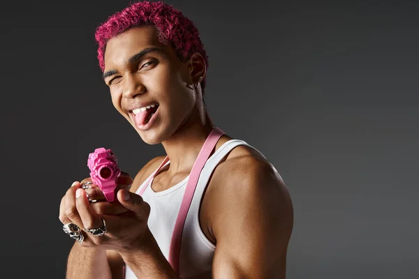 Juguetona modelo masculino de pelo rosa sobresaliendo de su lengua y nivelando su pistola de juguete rosa en la cámara - foto de stock
