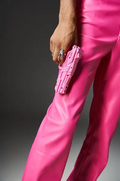 Vista recortada del hombre en pantalones de cuero rosa con anillo de plata sosteniendo pistola de juguete rosa sobre fondo gris - foto de stock
