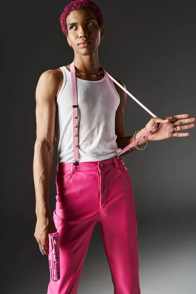 Gut aussehender Mann mit lockigem rosa Haar mit der Hand auf Hosenträgern, Spielzeugpistole haltend und wegschauend — Stockfoto