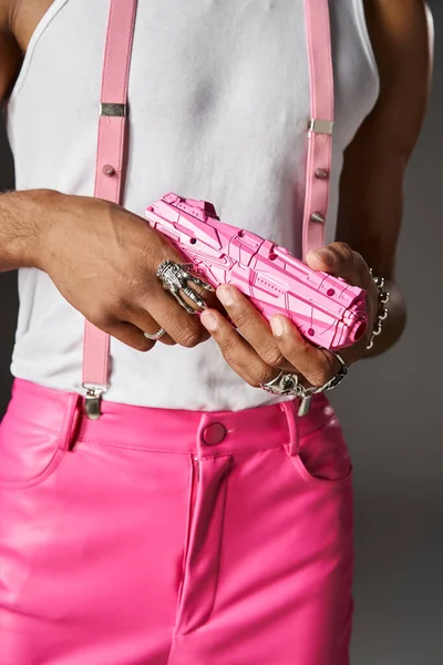 Ausgeschnittene Ansicht eines jungen männlichen Modells in rosa Hosen und Hosenträgern posiert mit Spielzeugpistole auf grauem Hintergrund — Stockfoto