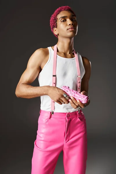 Homem de cabelos rosa elegante posando carismaticamente com arma de brinquedo no fundo cinza olhando para a câmera — Fotografia de Stock