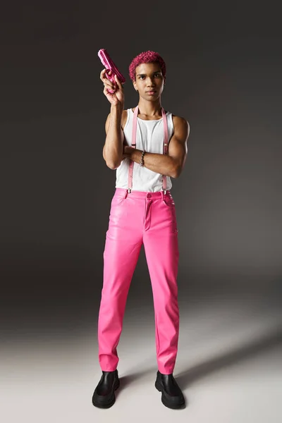Elegante hombre afroamericano con el pelo rosa apuntando a su pistola de juguete rosa posando sobre fondo gris - foto de stock