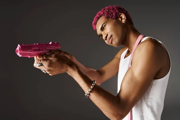 Rose cheveux afro-américain modèle masculin en pantalon avec des bretelles visant son pistolet jouet rose de côté — Photo de stock