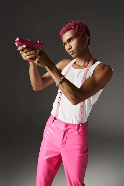 Hombre elegante guapo en pantalones de color rosa con tirantes con accesorios de plata con el objetivo de su pistola de juguete rosa - foto de stock