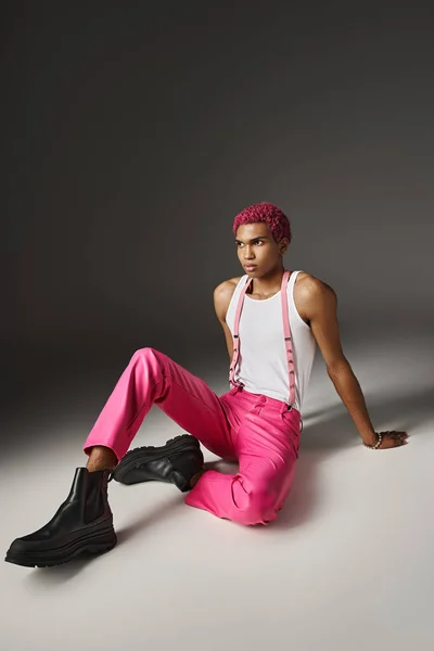 Bel homme aux cheveux roses en costume audacieux vif assis sur le sol et regardant ailleurs, concept de mode — Photo de stock