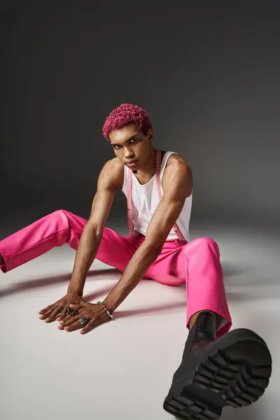 Stylish young man in pink pants with suspenders sitting on floor on gray backdrop, fashion concept — Stock Photo