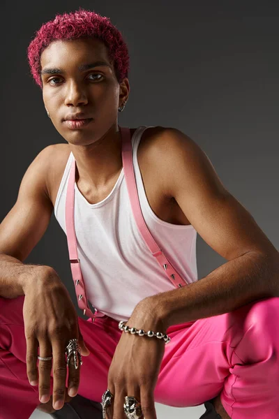 Молода стильна чоловіча модель з рожевим волоссям у яскравому вбранні, присідаючи на сірому фоні, концепція моди — стокове фото