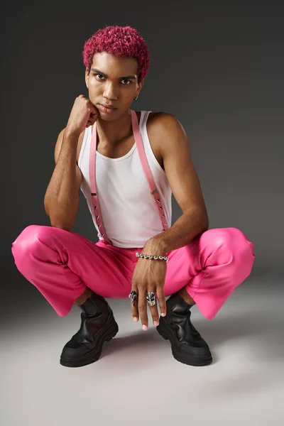 Trendy african american man posing on gray backdrop squatting and looking at camera, fashion concept — Stock Photo