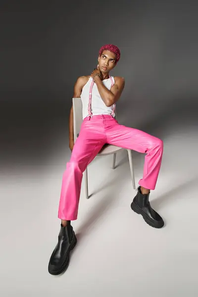 Modèle masculin afro-américain en pantalon rose avec bretelles posant sur chaise blanche, concept de mode — Photo de stock