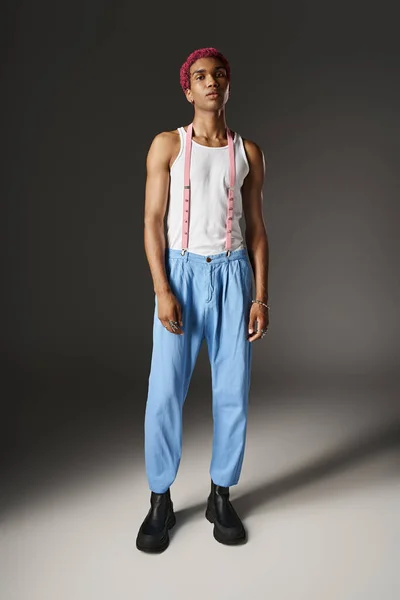 Handsome man with pink hair on gray backdrop in blue pants with pink suspenders, fashion concept — Stock Photo