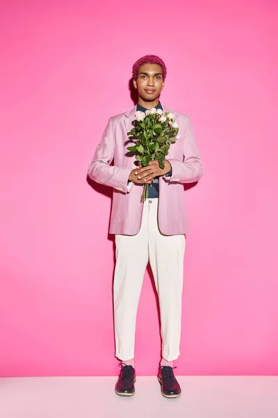 Homem de cabelos cor-de-rosa bonito posando com buquê de rosa no fundo rosa e sorrindo alegremente — Fotografia de Stock