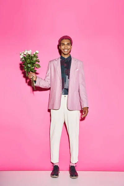 Весёлый молодой человек с букетом белых роз, позирующих на розовом фоне, искренне улыбающийся в камеру — стоковое фото