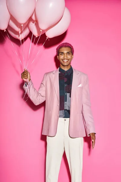 Cheerful male model smiling happilly with balloons in hands and looking at camera on pink backdrop — Stock Photo