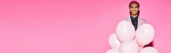 Jovem bonito sorrindo anormalmente com balões em mãos posando no fundo rosa, banner — Fotografia de Stock