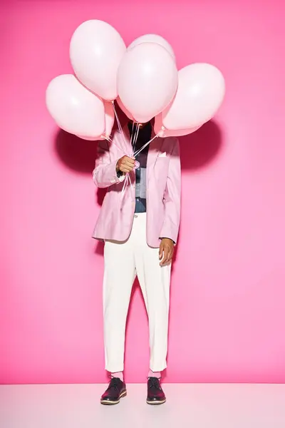 Homme élégant en tenue vibrante tenant des ballons devant son visage posant sur fond rose — Photo de stock