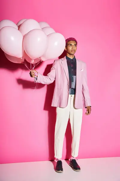 Bel homme aux cheveux rose bouclés en blazer rose posant avec des ballons sur fond rose, poupée comme — Photo de stock