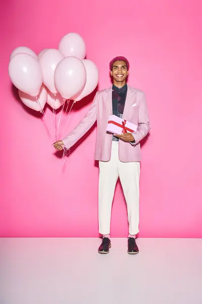 Joyous young man in blazer posing with balloons and present smiling unnaturally on pink backdrop — Stock Photo