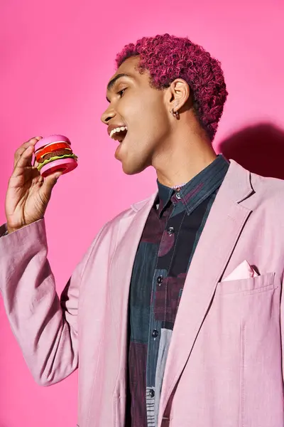 Allegro giovane uomo in giacca rosa con orecchini d'argento mangiare mini hamburger posa su sfondo rosa — Foto stock
