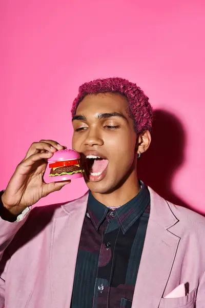 Stylish good looking man in pink blazer posing unnaturally eating mini burger on pink bakcground — Stock Photo