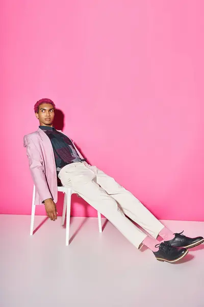Young handsome man in pink blazer with white pants sitting on chair on pink backdrop, doll like — Stock Photo