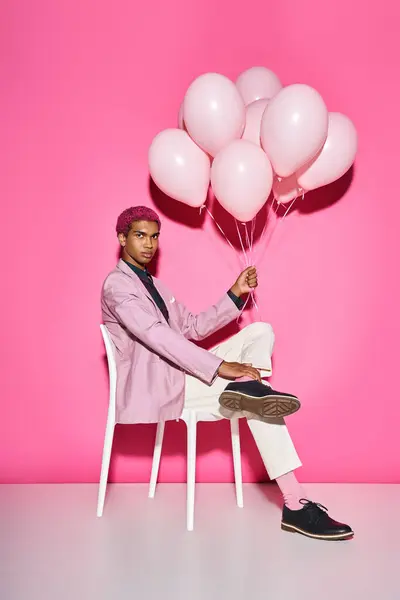 Homem de cabelos rosa bonito sentado na cadeira branca e segurando balões olhando para a câmera — Fotografia de Stock