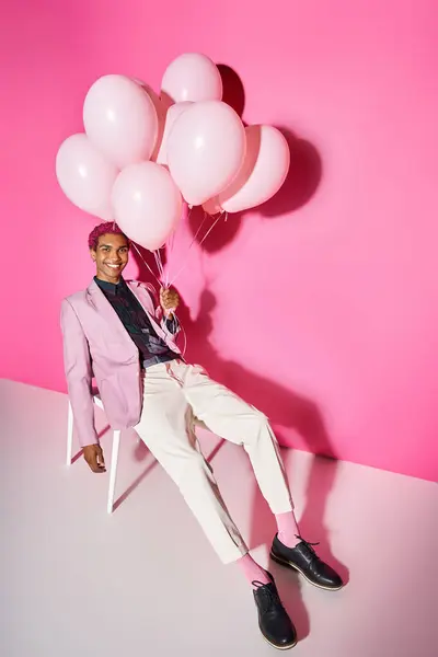 Jovem alegre em blazer rosa posando na cadeira branca com balões na mão, agindo como boneca masculina — Fotografia de Stock