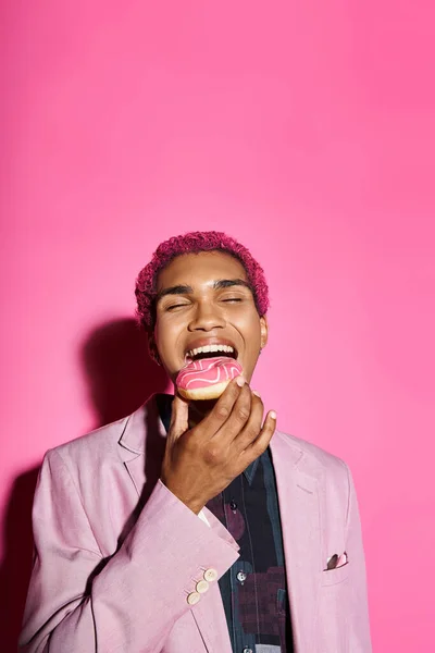 Homem elegante bonito com cabelo rosa encaracolado em blazer elegante comer donut posando em pano de fundo rosa — Fotografia de Stock