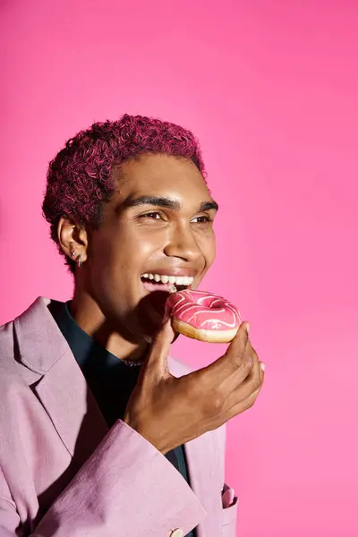 Joyeux jeune homme afro-américain en blazer rose et boucles d'oreilles profiter de son beignet sur fond rose — Photo de stock