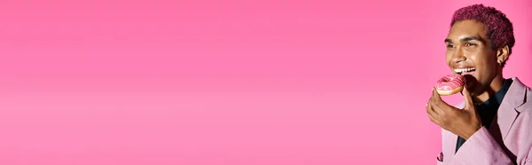 Joyous young man with curly hair and earrings enjoying donut posing on pink background, banner — Stock Photo