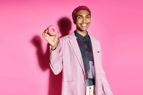 Homme joyeux en tenue vibrante souriant anormalement à la caméra et tenant beignet sur fond rose — Photo de stock