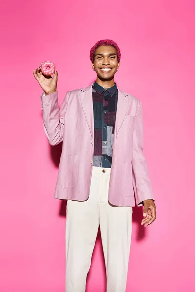 Bello africano americano maschio modello posa innaturalmente sorridente e tenendo ciambella, rosa sfondo — Foto stock