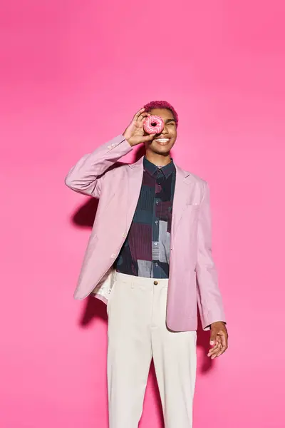 Игривый молодой человек с кудрявыми розовыми волосами позирует с пончиком рядом с лицом, позируя на розовом фоне — стоковое фото