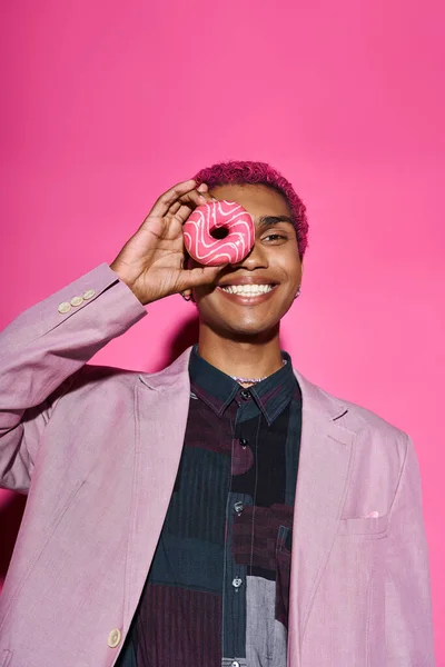 Modelo masculino de boa aparência em traje vibrante posando de forma não natural com donut perto do rosto em pano de fundo rosa — Fotografia de Stock