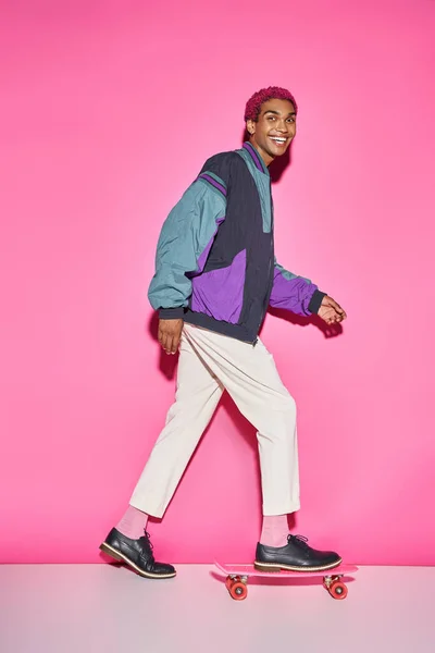 Joyeux jeune modèle masculin posant sur skateboard dans le profil et souriant à la caméra sur fond rose — Photo de stock