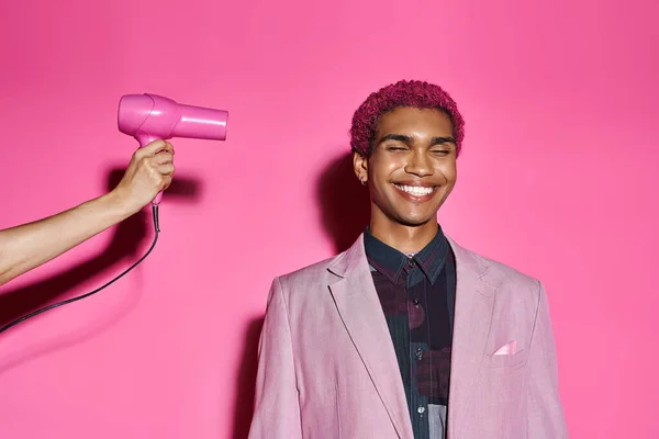 Homem bonito com cabelo rosa encaracolado posando com olhos fechados em rosa back drop, mão com secador de cabelo — Fotografia de Stock