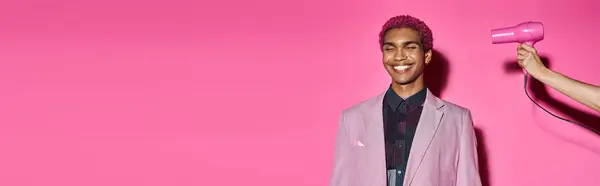 Homem sorridente posando de forma não natural no fundo rosa com olhos fechados, mão com secador de cabelo, banner — Fotografia de Stock